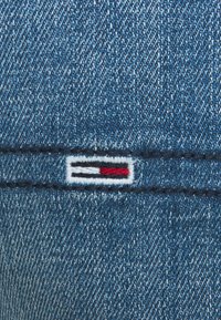Blue denim fabric featuring a white stitched label with a horizontal pattern of red, white, and black stripes. Texture is smooth and textural.