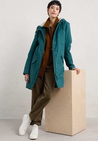 Teal vettävä parka, jonka huppu on vuorattu turkiksella, ruskean corduroy-takin päällä, yhdistettynä oliivinvihreisiin housuihin ja valkoisiin lenkkareihin.