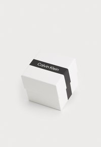 Boîte-cadeau blanche avec un bandeau noir étiqueté "Calvin Klein". Forme carrée avec une finition lisse et mate et un couvercle plier en toute sécurité.
