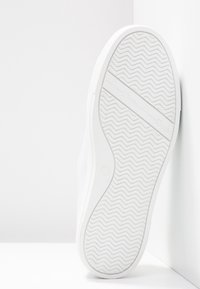 Semelle blanche de basket avec un motif ondulé et une bande horizontale affichant un texte en relief discret sur fond blanc.