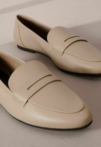Mocassini in pelle beige con una finitura liscia, caratterizzati da una decorativa cucitura in alto e dal design slip-on, poggiati su una suola bassa in gomma nera.