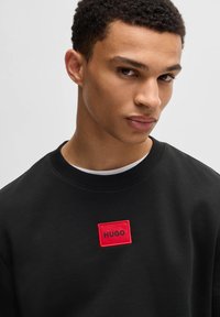 Svart sweatshirt i bomull med en röd broderad logotyp som säger "HUGO" på framsidan. Rundhalsad design.
