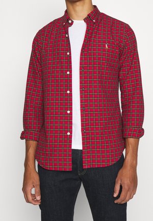 Homme portant une chemise à carreaux rouge et verte à manches longues par-dessus un t-shirt blanc et un jean foncé, avec les manches retroussées jusqu'aux coudes.