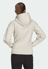 Krémová mikina s velkou kapucí, žebrovanými akcenty na ramenou a přiléhavými rukávy. Zadní strana má hladkou texturu a minimální švy.