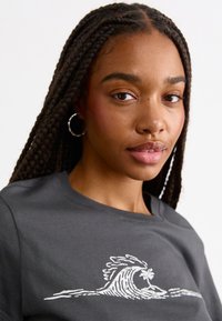 Dark grey t-shirt featuring a white wave graphic design. The fabric appears smooth and has a crew neckline. Braided hair and hoop earrings visible.