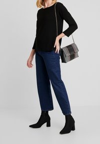 Schwarzes Langarmoberteil, blaue High-Waist-Jeans und schwarze Stiefeletten mit Blockabsätzen. Schwarz-weiße karierte Handtasche mit Kettenriemen.