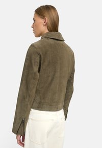 Olive zielona kurtka z zamszu z wyciętym kołnierzem, skróconym fasonem i suwakami na mankietach, o miękkim, teksturowanym wykończeniu i minimalnym przeszyciu.