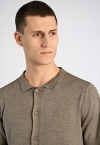 Brunstickad polo med krage, utrustad med tre bruna knappar och en diskret logo broderad på bröstet. Korta ärmar.