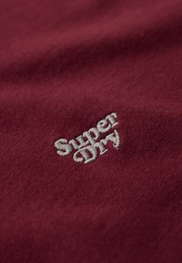 Wit geborduurd "Super Dry" logo op een gestructureerde kastanjebruine stof, die de weving van de stof en lichte kreukels toont.