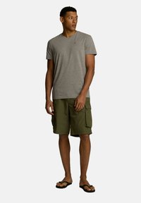 Szary t-shirt w serek, oliwkowe szorty cargo z kieszeniami bocznymi oraz czarne klapki. Swobodny letni strój zaprezentowany na modelu.