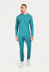 Türkisfarbenes langärmeliges Oberteil und passende Hose aus glattem, dehnbarem Stoff mit elastischen Bündchen und lockerem Schnitt. Weiße Sneaker ergänzen das Outfit.