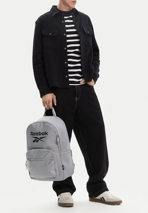 Hombre con chaqueta negra, camisa de rayas blancas y negras, y pantalones negros sostiene una mochila gris de Reebok al lado. Zapatillas blancas completan el conjunto.