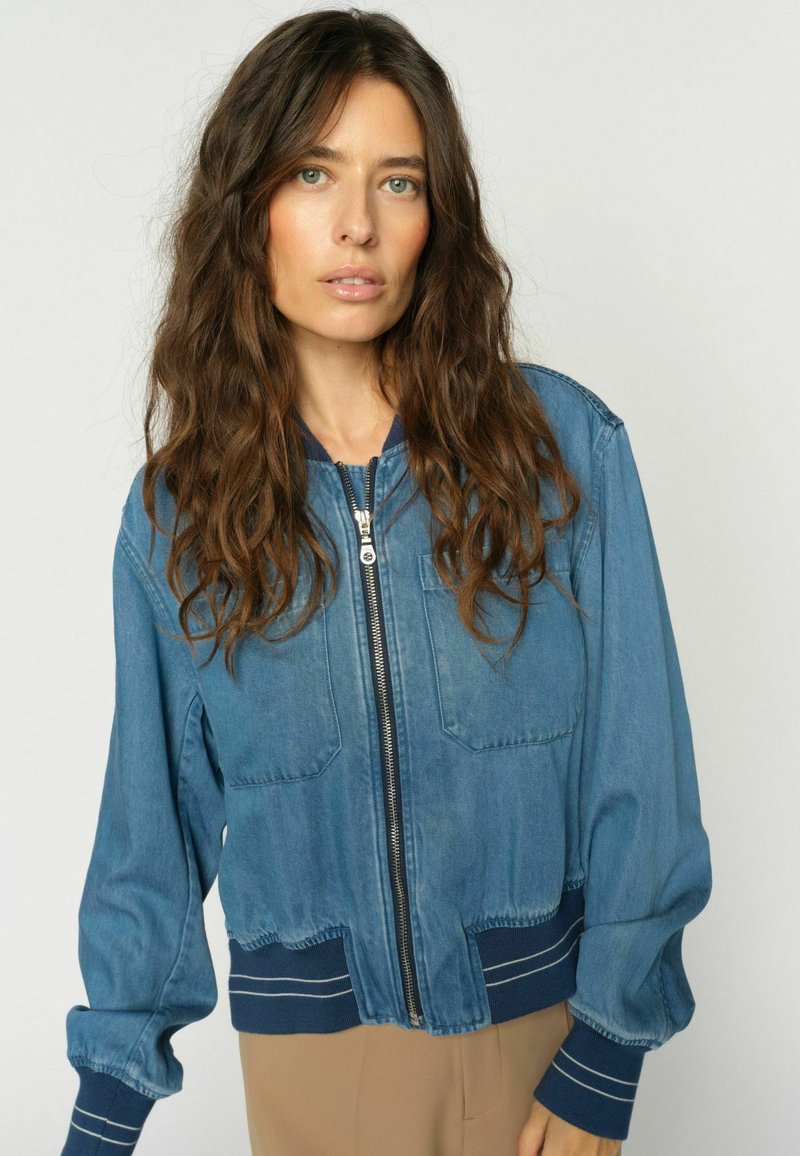 Femme aux longs cheveux bruns ondulés portant une veste en denim bleue à fermeture éclair avec des poignets côtelés et un pantalon beige, debout devant un fond uni.