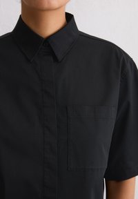 Camicia nera a maniche corte con colletto a punta e taschino sul petto. Realizzata in tessuto morbido con un design pulito e minimalista.