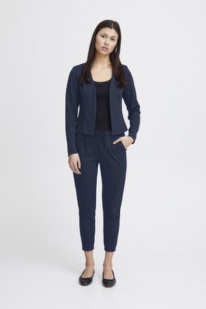 Ženska stoji oblečena v mornarsko modro suknjič, ujemajoče se hlače z vrvico, črno majico in črne balerinke na preprostem belem ozadju.