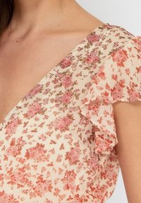 Kukallinen mekko kevyestä kermanvärisestä kankaasta, jossa on vaaleanpunaisia ja rustisia kukkakuoseja. Varustettu röyhelöhihoilla ja V-pääntiellä.