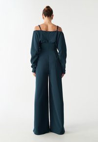 Tumši tirkīza krāsas kombinezons ar plašām kājām, bez plecu siksnām, garām piedurknēm un pieguļošu vidukli. Gluda auduma ar maigu spīdumu. Aizmugurējā rāvējslēdzēju aizdare.