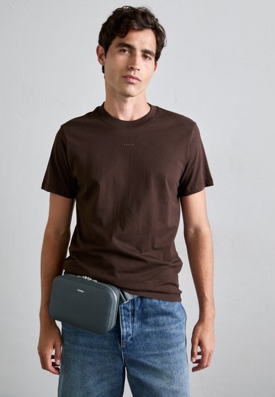 Brun bomull t-skjorte med korte ermer, kombinert med lyseblå denimjeans. En grå rektangulær crossover-veske hviler ved livet.