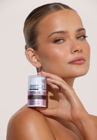 Siero abbronzante in una bottiglia trasparente con pompa e etichetta rosa, caratterizzato da una fascia argentata. La modella lo tiene vicino al viso, mostrando una pelle liscia.