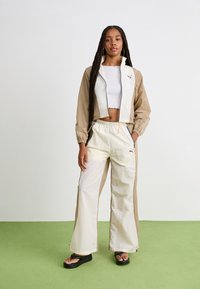 Chaqueta cropped beige y blanca con cremalleras, combinada con pantalones blancos que tienen paneles laterales beige. Llevada con sandalias de plataforma negras.