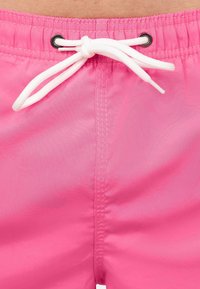 Shorts de bain roses en matériau léger avec une taille élastique, dotés d'un cordon blanc et d'œillets noirs pour l'ajustement.