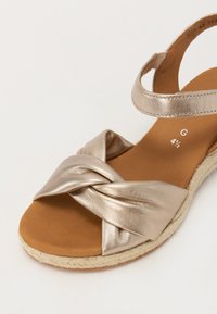 Metallic gold sandal with a knotted front design, woven jute sole, and adjustable ankle strap, featuring a smooth finish and soft texture.