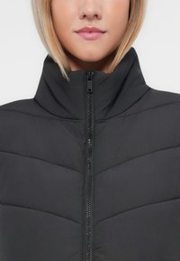 Zwarte pufferjack met hoge kraag, met geborduurde chevron details en een ritssluiting aan de voorkant. Glad, gewatteerd textuur.