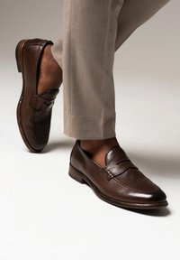 Braune Lederloafers mit glatter Textur, versehen mit einer genähten Akzentuierung an der Vorderseite und einem niedrigen Absatz, kombiniert mit hellgrauen Hosen.