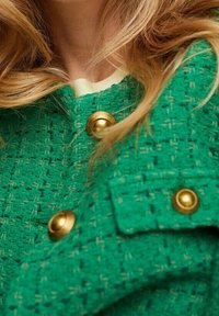 Giacca in tweed verde con una finitura texture. Presenta bottoni in tonalità oro e un dettaglio del colletto crema a contrasto. Vista ravvicinata.