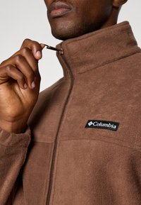 Smeđa flis jakna s visokim ovratnikom, zatvaranjem na zip i Columbia logotipom na lijevoj strani prsiju. Mekana tekstura, ležeran dizajn pogodan za slojevito nošenje.