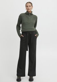 Pull en col roulé en tricot vert olive avec une fermeture éclair quart, associé à un pantalon noir à taille haute et large et des bottes noires chunky.