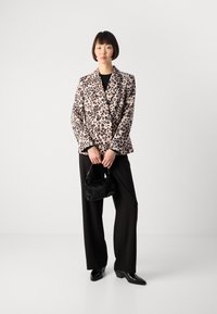 Vrouw in een blazer met luipaardprint, zwarte wijde broek, zwarte enkellaarsjes, met een kleine zwarte handtas tegen een effen witte achtergrond.