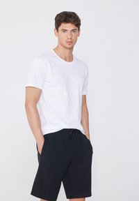 Camiseta de algodón blanca con mangas cortas y un bolsillo en el pecho, combinada con pantalones cortos negros con cordón. Diseño simple y corte relajado.