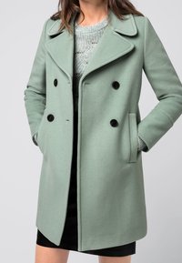 Svetle zelený, dvouřadový vlněný kabát s límcem; má černé knoflíky, boční kapsy a rovný střih. Tkanina se zdá být hladká a texturovaná.