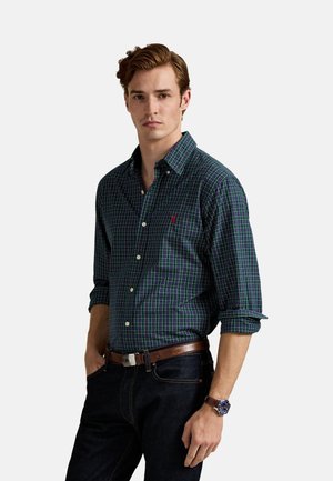 Camisa a cuadros en azul marino y verde con mangas largas, cuello abotonado y un pequeño logo. Se lleva con jeans oscuros y un cinturón de cuero marrón.