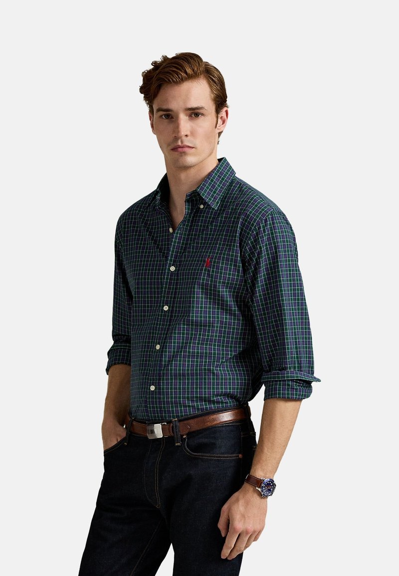 Camisa a cuadros en azul marino y verde con mangas largas, cuello abotonado y un pequeño logo. Se lleva con jeans oscuros y un cinturón de cuero marrón.