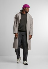 Cappotto lungo grigio sopra un hoodie nero, abbinato a jeans scuri e sneakers multicolori. L'individuo indossa un berretto di lana viola brillante.