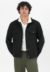 Chaqueta negra y texturizada con dos bolsillos en el pecho y botones a presión. Presenta una camiseta interior con cuello blanco, combinada con pantalones de color verde oliva.