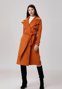 Trench-coat en mélange de laine orange avec de grands revers, taille ceinturée et poches latérales ; associé à des bottes noires en daim montantes.