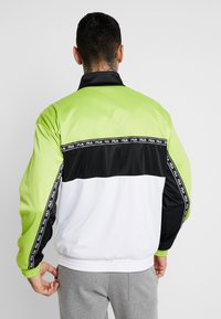 Chaqueta cortavientos colorida con secciones en verde lima, negro y blanco, presenta una banda con el logo de Fila y puños y dobladillo acanalados.
