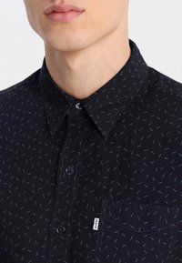 Svart skjorta med ett fint, spritt blått mönster, knappad krage, bröstficka och varumärkesetikett på fickan.