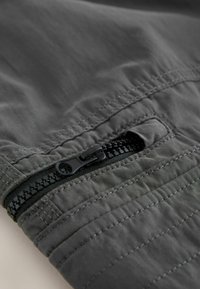 Gros plan d'une fermeture éclair noire sur un vêtement en tissu gris foncé avec des coutures détaillées et un curseur de fermeture partiellement ouvert.
