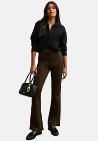Sweat à zip noir, pantalon évasé en imprimé léopard marron, sac à main noir avec charme, et chaussures noires avec des détails texturés.