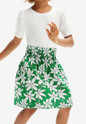 Barn iført en hvid kortærmet skjorte og et grønt nederdel med hvidt blomstermønster, med den ene hånd hævet.
