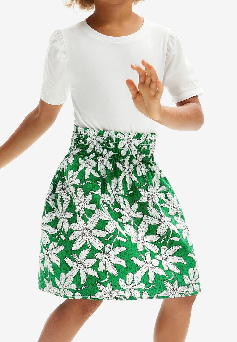 Enfant portant une chemise blanche à manches courtes et une jupe verte à motif floral blanc, avec une main levée.