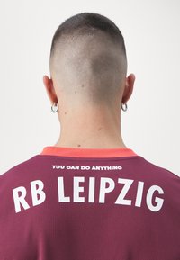 Maglia bordeaux con maniche corte, caratterizzata dalla scritta "RB Leipzig" in bianco sulla schiena. Tra i dettagli, un colletto rosa e un tessuto testurizzato.