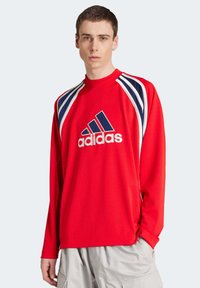 T-shirt athlétique rouge à manches longues avec un col rond, présentant des accents rayés bleu marine et blanc et un grand logo Adidas blanc à l'avant.