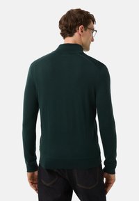 Mann in einem dunkelgrünen Langarmpullover und dunklen Jeans, von hinten vor einem schlichten hellen Hintergrund abgebildet.