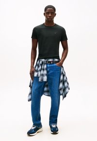 Uomo che indossa una maglietta nera, jeans blu, sneaker blu navy e una camicia a quadri bianca e nera legata in vita, in piedi con una mano in tasca.