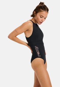 Maillot de bain noir une pièce avec un col montant, des découpes sur les côtés et une fermeture éclair. Présente un logo rose sur le côté gauche. Tissu lisse et extensible.
